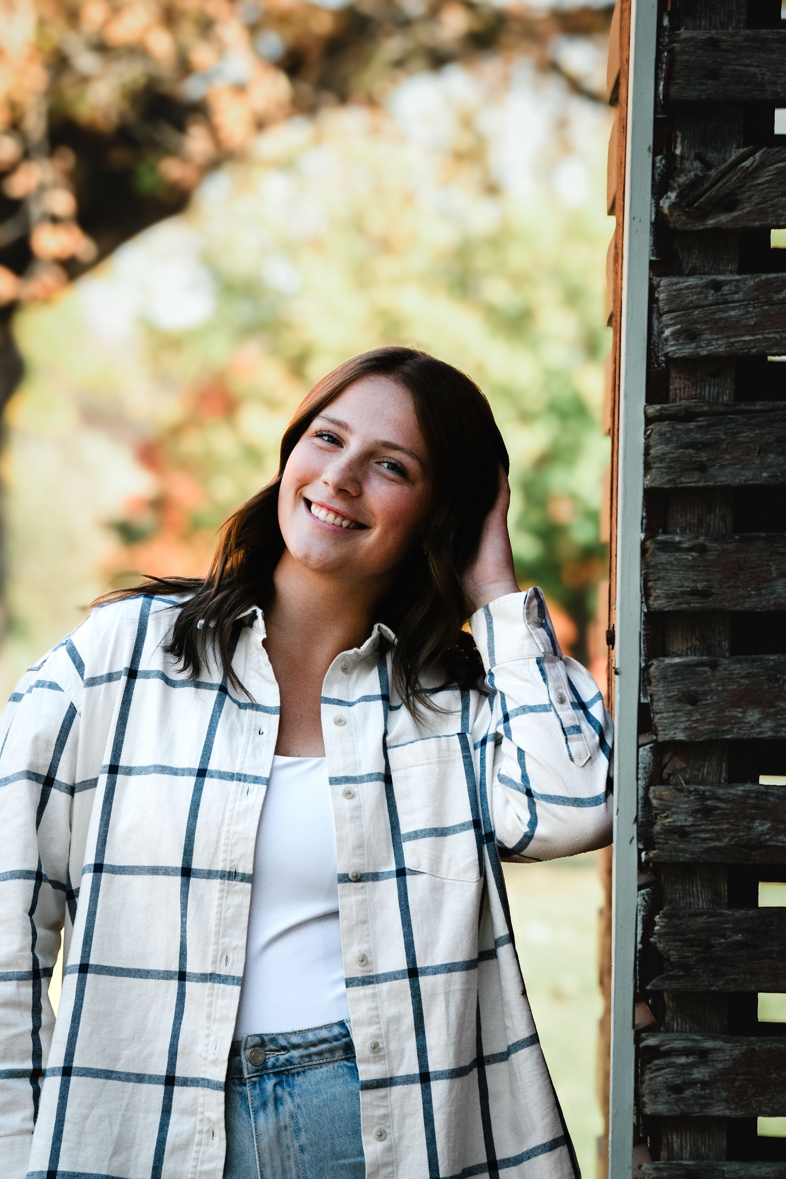 Smiling young woman in plaid shirt leaning against rustic wooden barn door with autumn foliage background.