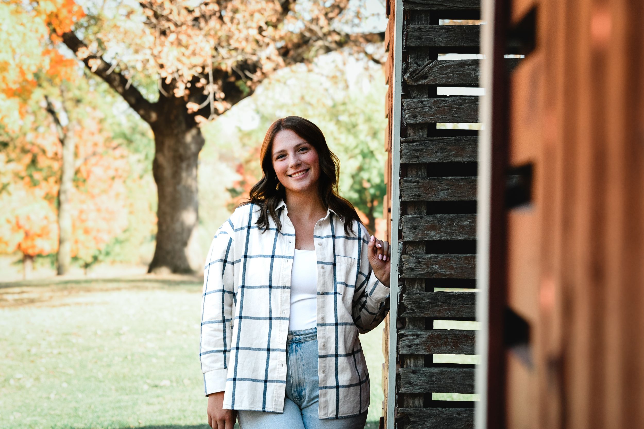 Woman in plaid shirt smiling outdoors near rustic barn with colorful autumn trees in background.