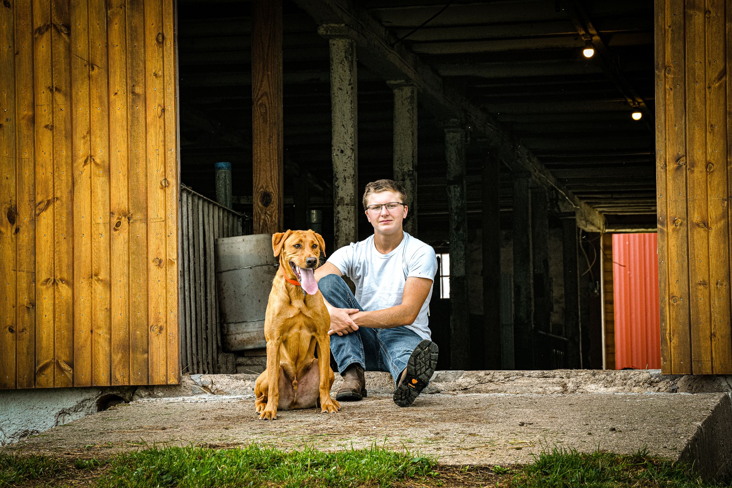 Man crouching beside a brown Vizsla dog in front of a rustic wooden barn entrance.