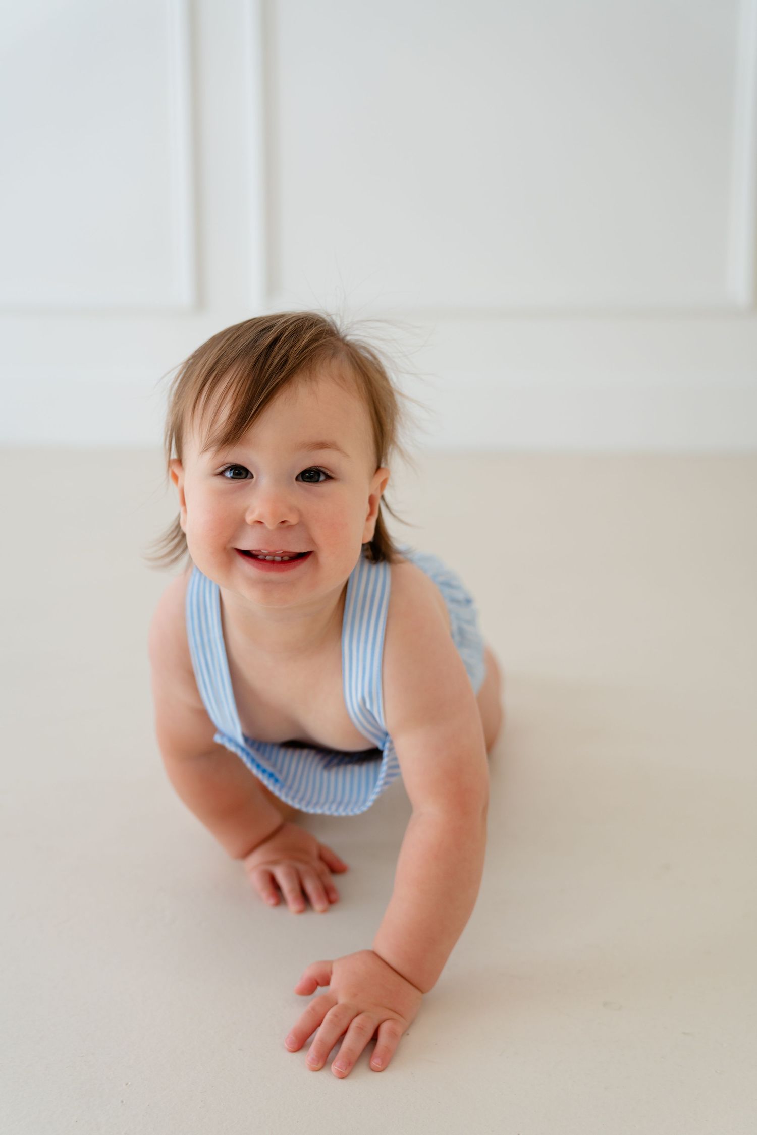 A happy baby in a blue striped romper crawling on a white floor, smiling at the camera.