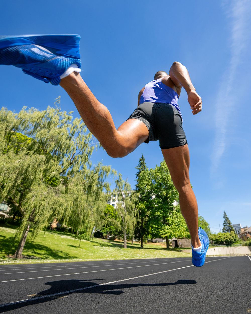 Low-angle view of an athlete sprinting on a track in blue shoes and shorts under a bright blue sky.