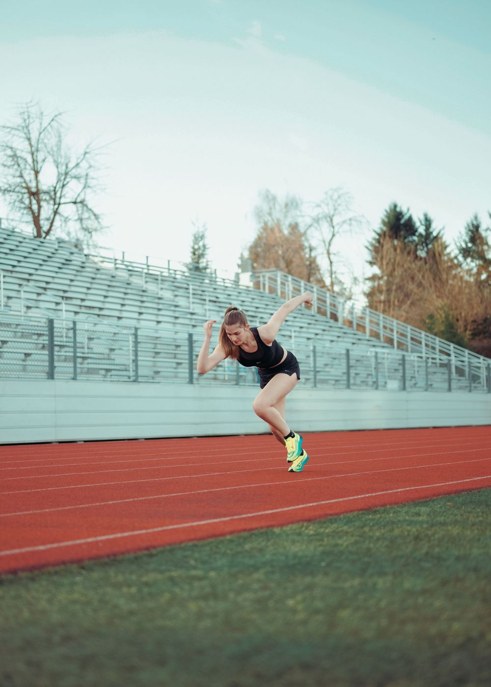 Female track athlete sprinting on red running track with empty bleachers and trees in background.