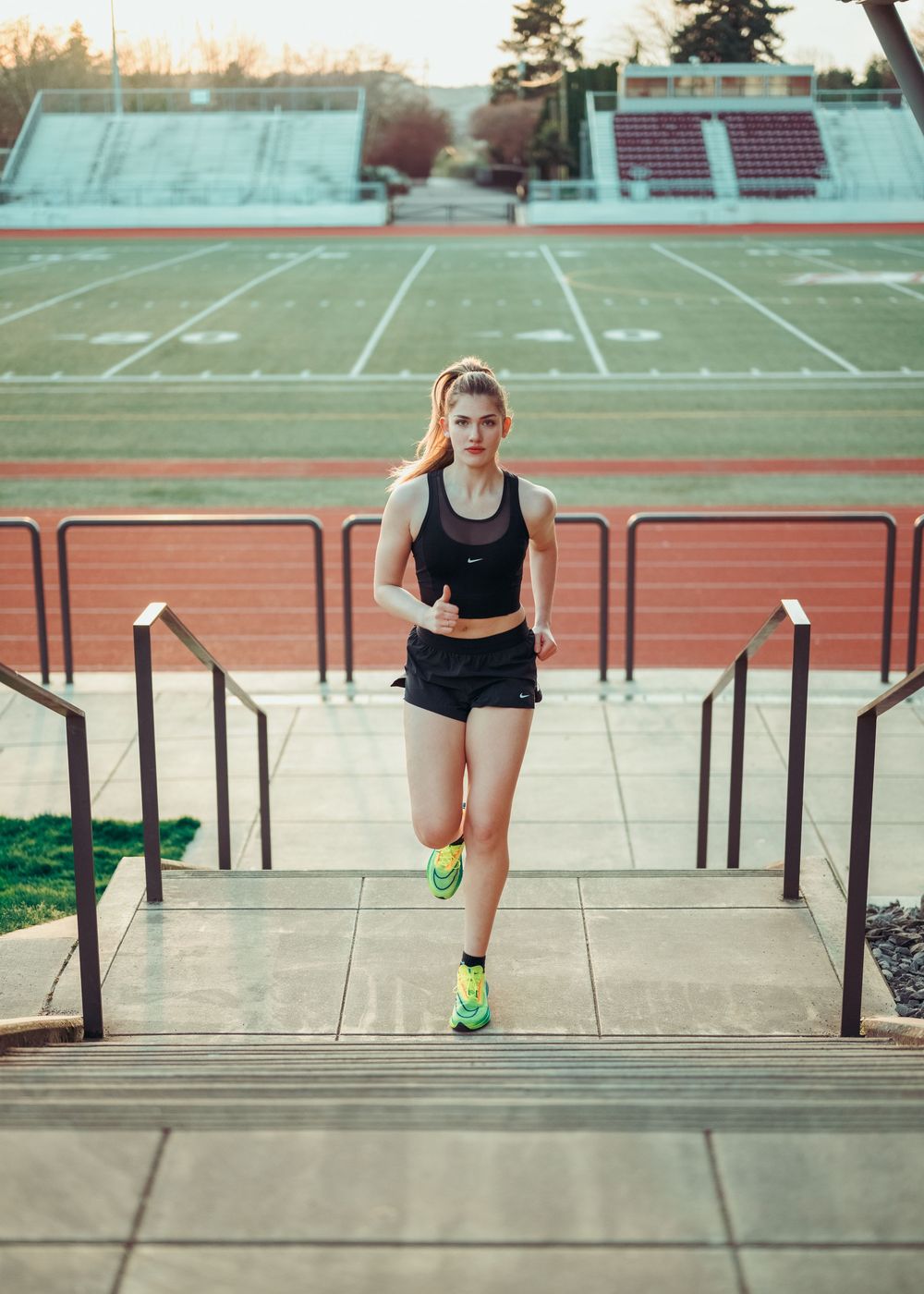 A young female athlete in black sportswear and neon shoes runs up stadium stairs near a track and field.