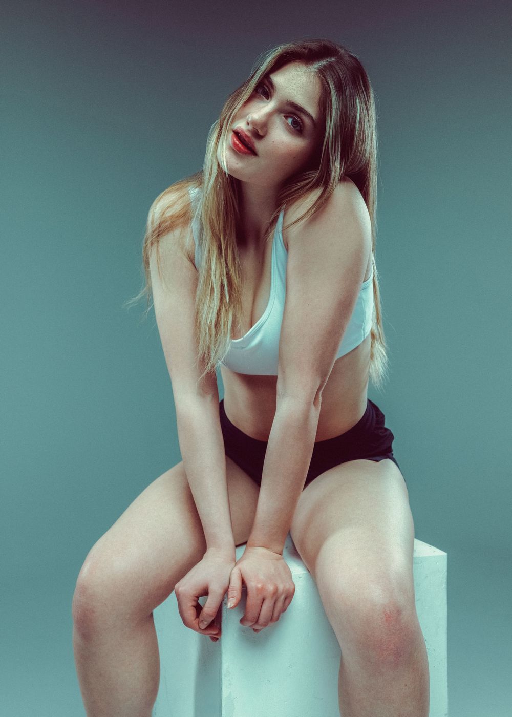 A young woman with long blonde hair poses seated on a white cube in a light blue sports bra and black shorts.