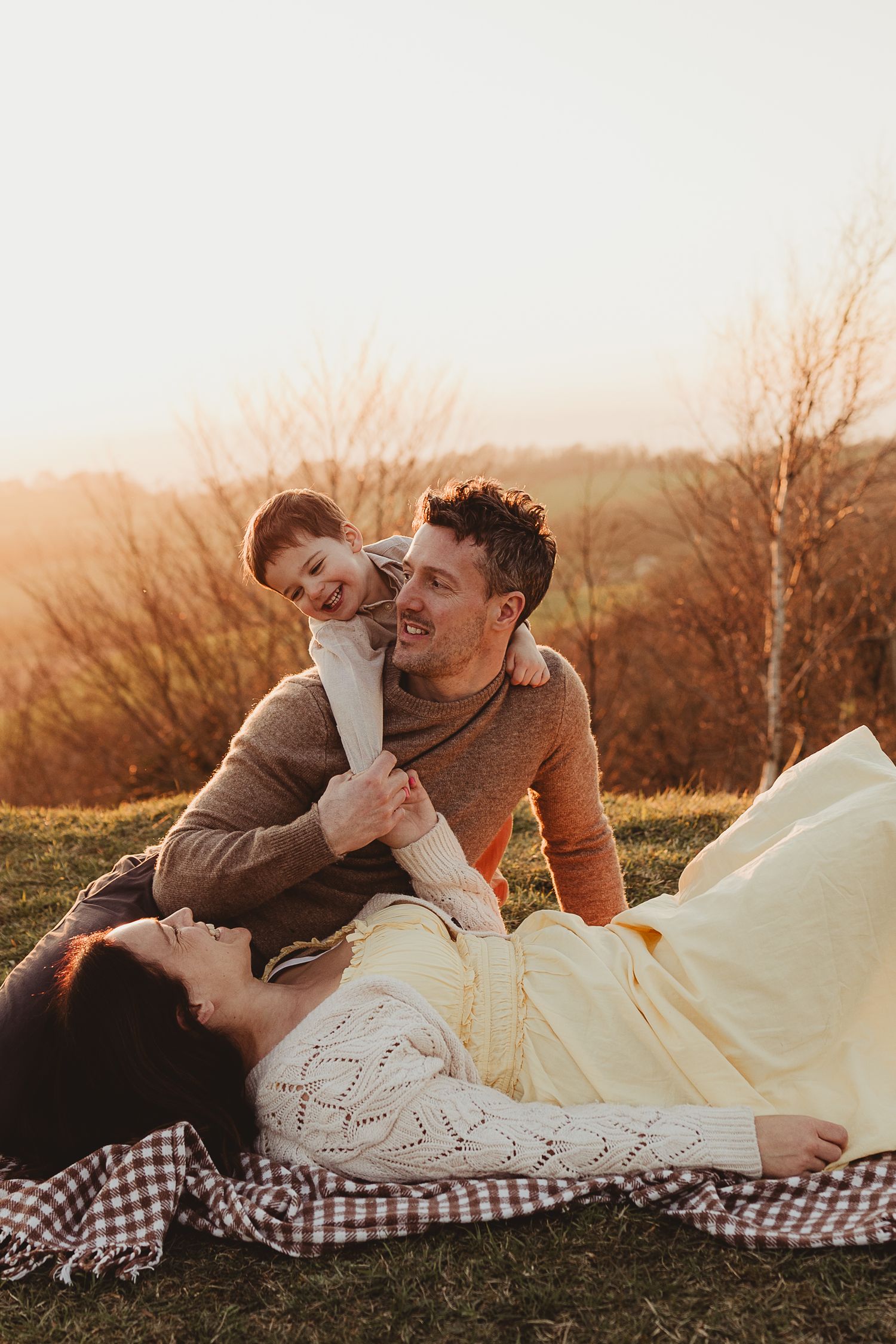 Family relaxing on a grassy hill at golden hour, with a toddler playing beside reclining parents.