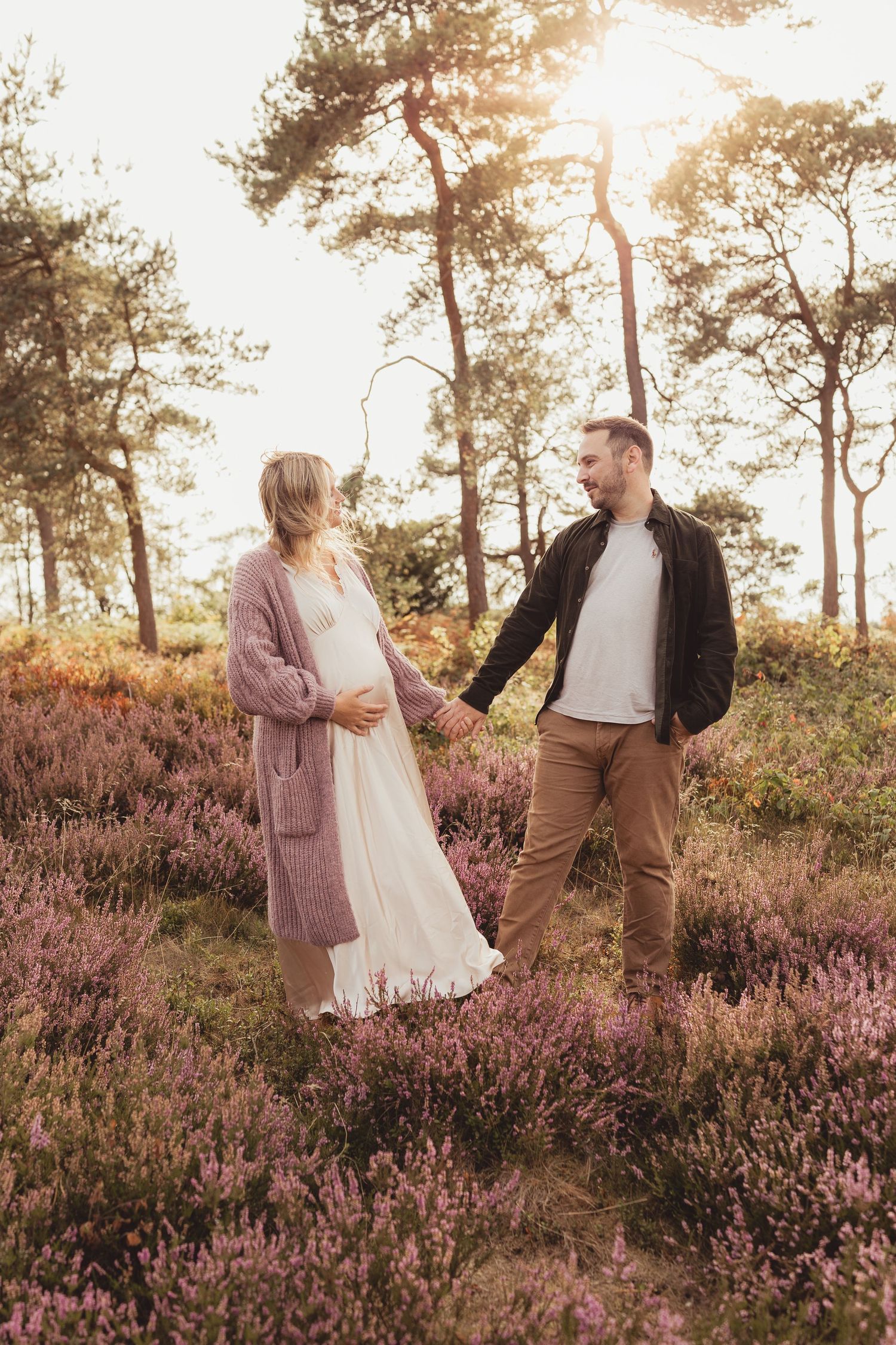 Pregnant couple holding hands in blooming heather field at golden hour, surrounded by pine trees.