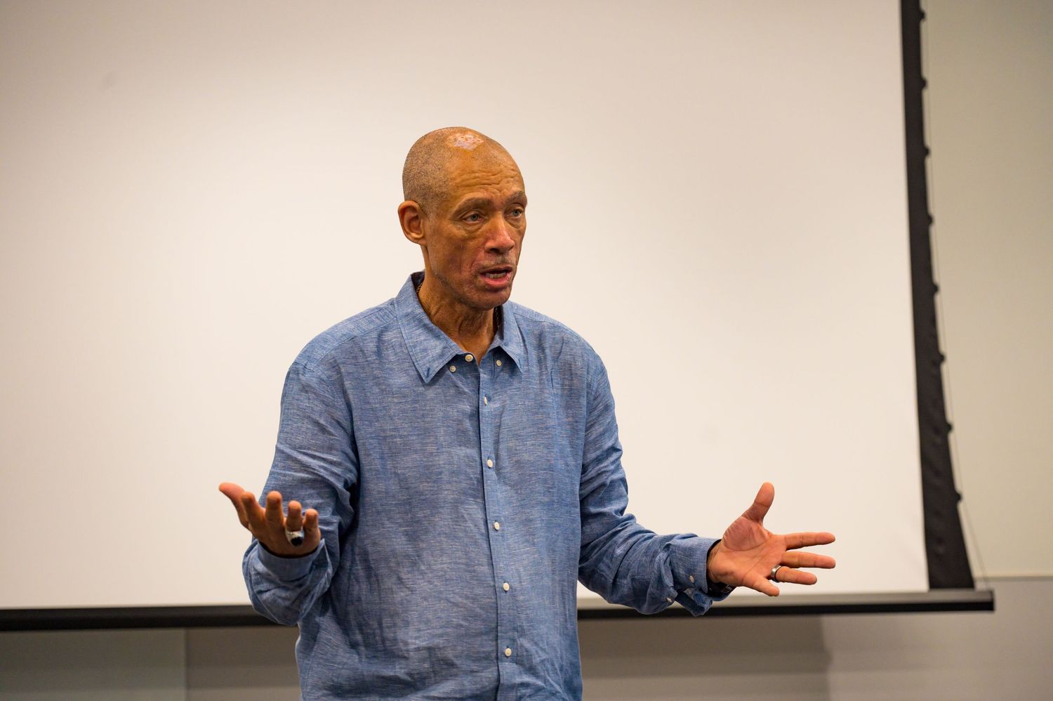 Speaker in blue shirt presenting with open hand gestures in a classroom setting.