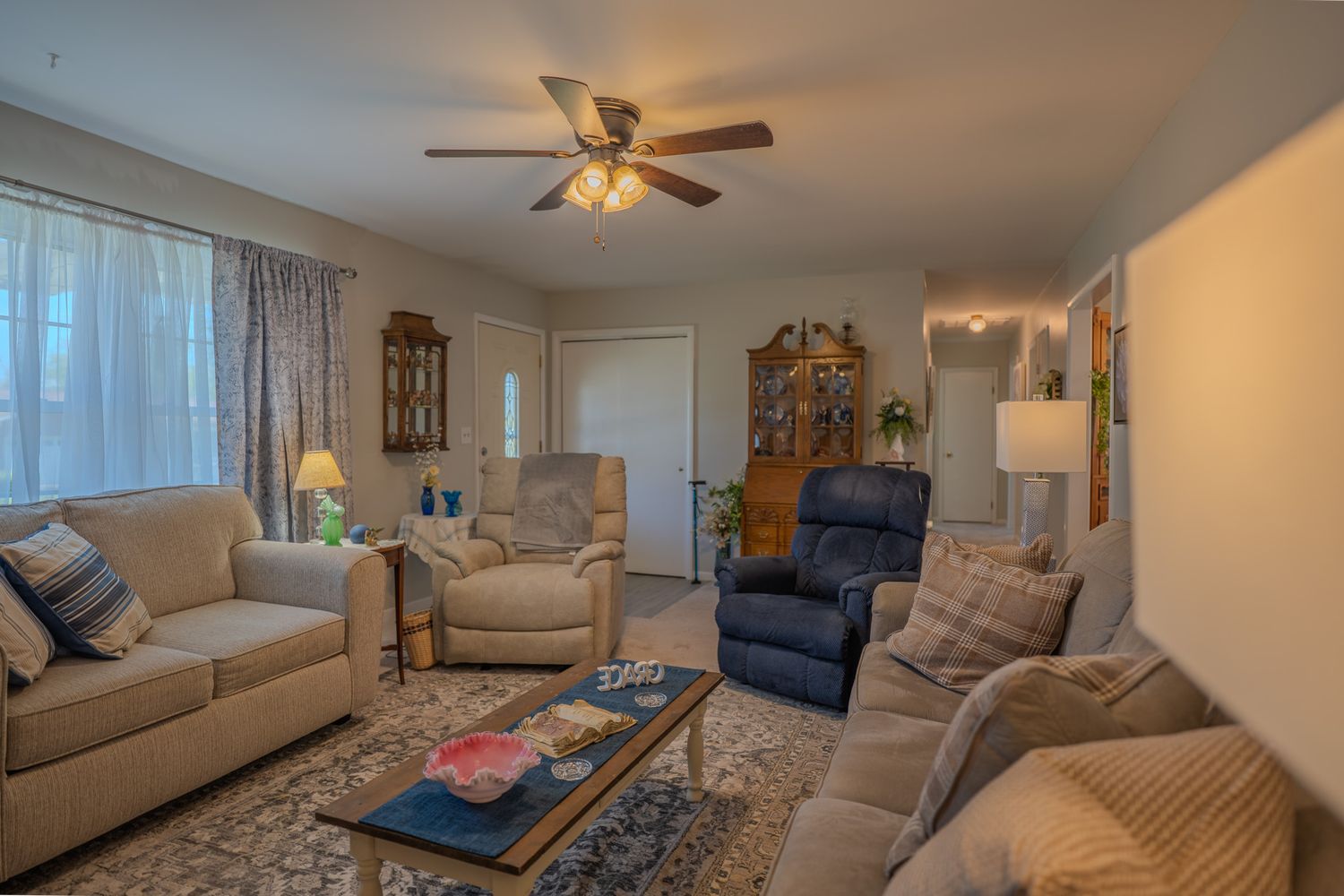 Cozy living room with beige sofas, recliner chair, ceiling fan, wood furniture, and natural light from large windows.