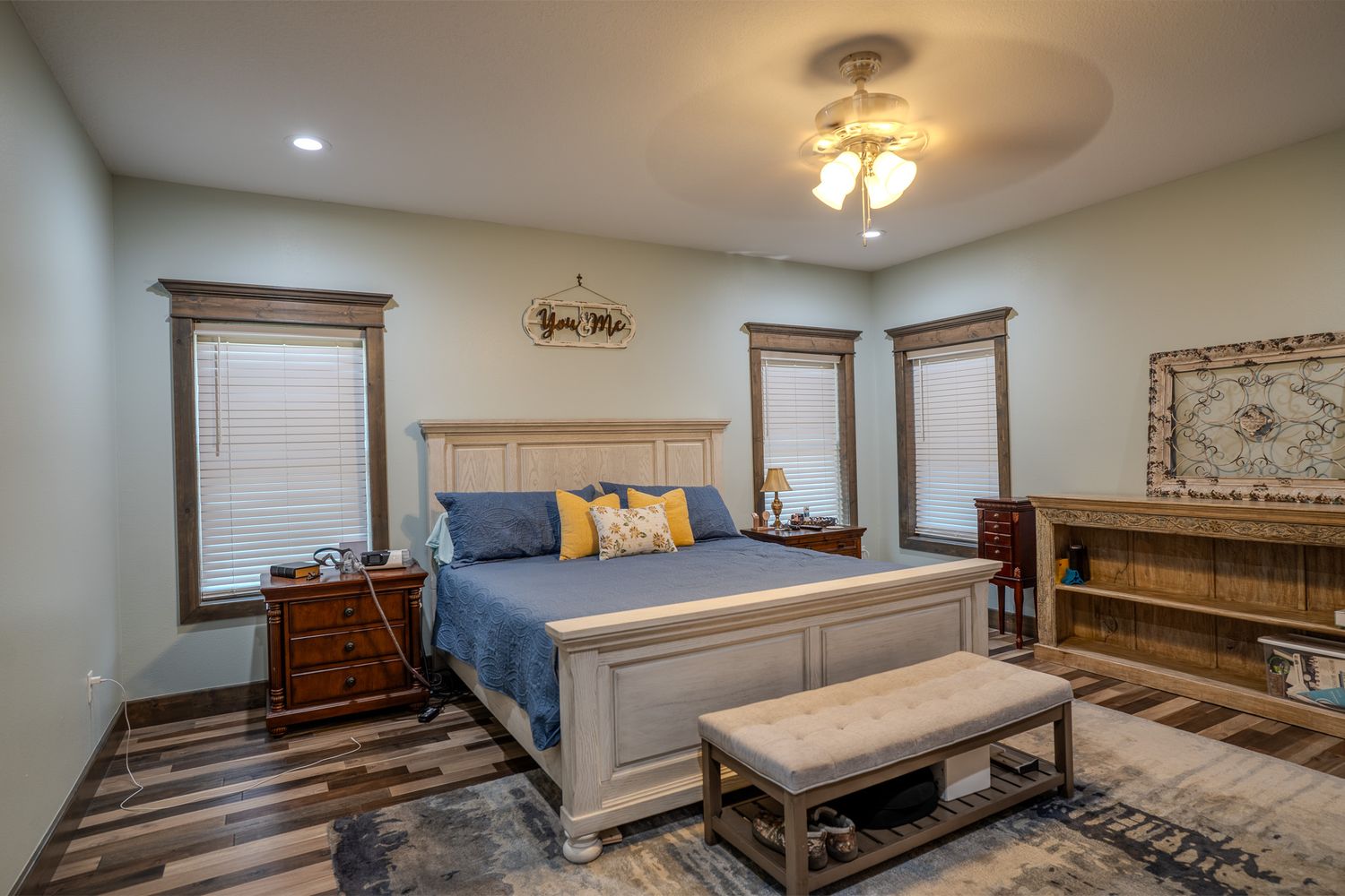 Spacious master bedroom with white bed frame, blue bedding, hardwood floors, ceiling fan, and two windows with blinds.