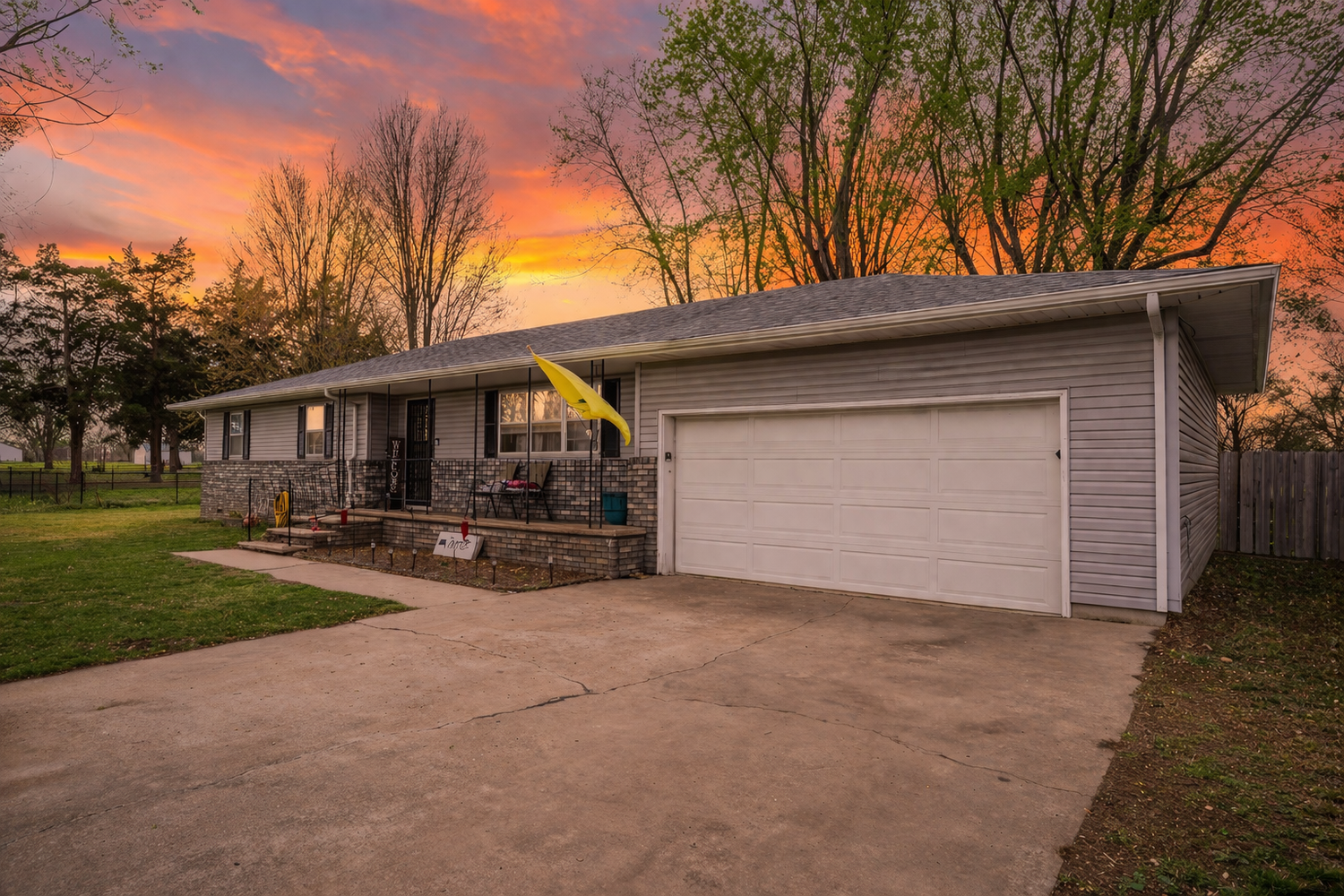 Ranch-style home with two-car garage at sunset, featuring stone accents and a covered porch amid lush green trees.
