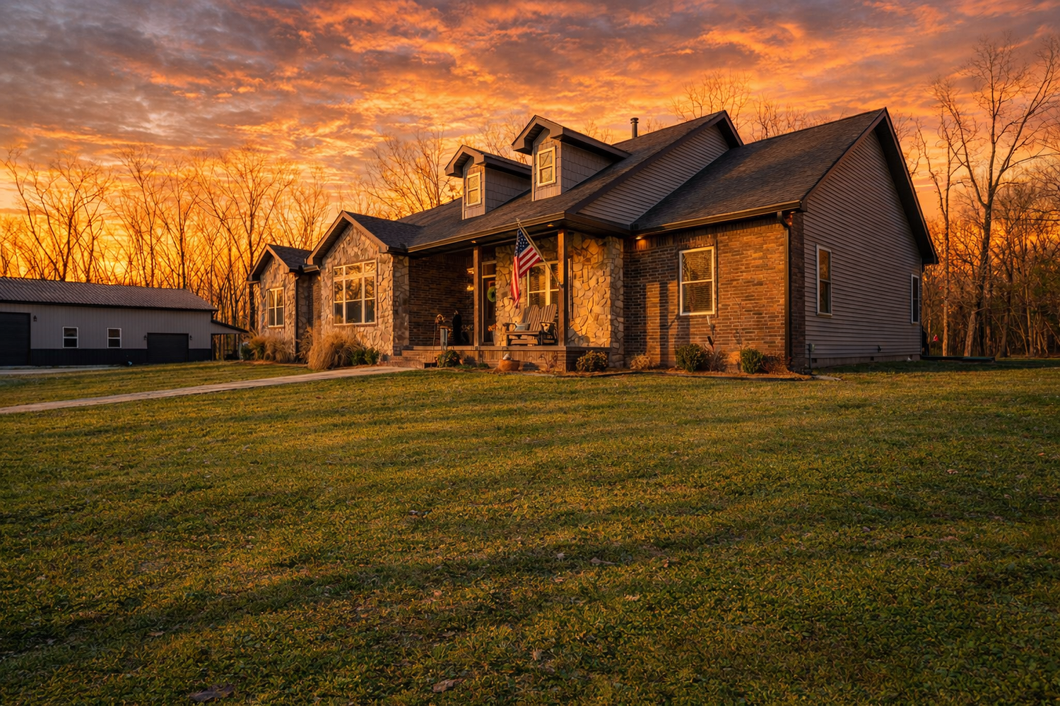 Stone farmhouse with American flag glows under dramatic orange sunset sky, surrounded by lush green lawn and bare winter trees.