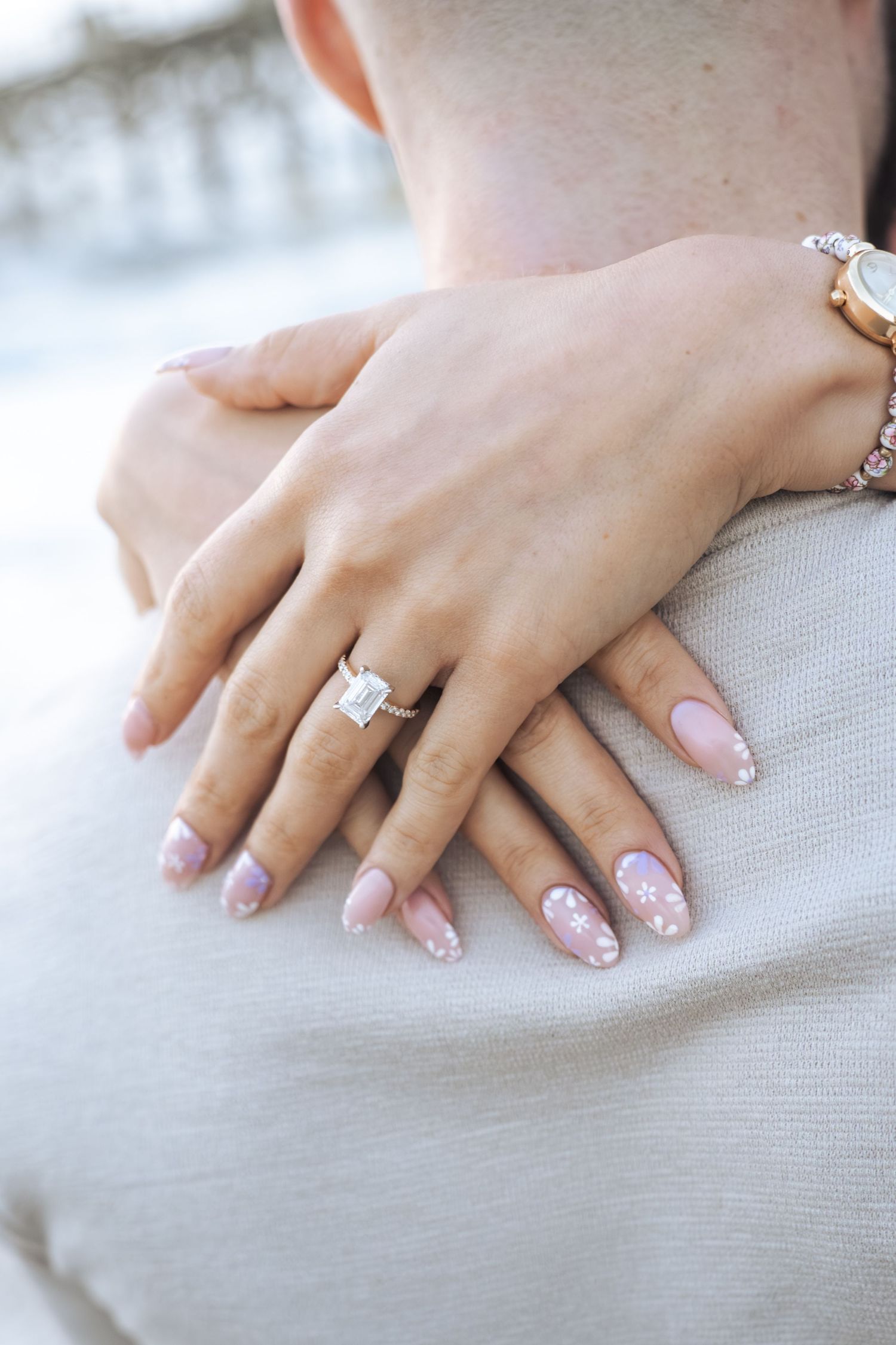 Close-up of a woman's hand with pink floral nail art and diamond engagement ring resting on a man's shoulder.