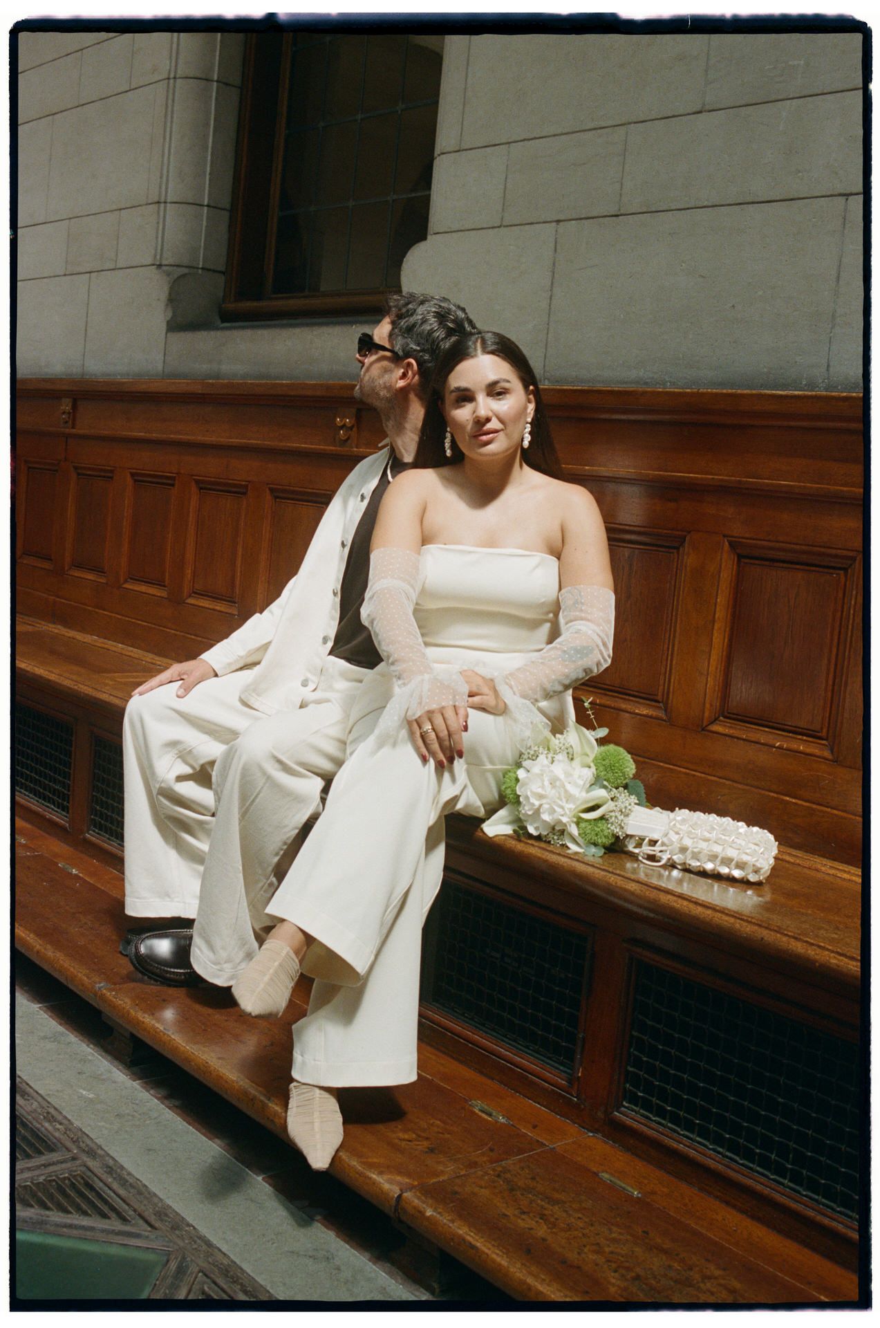 Bride in white suit and groom in black tuxedo pose in a modern venue with wood-paneled walls and warm lighting.