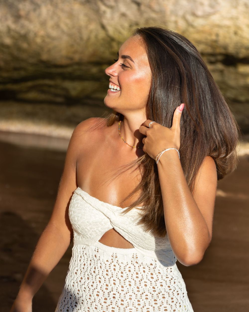 Smiling brunette woman in white crochet dress poses at beach with rocky backdrop in golden sunlight.