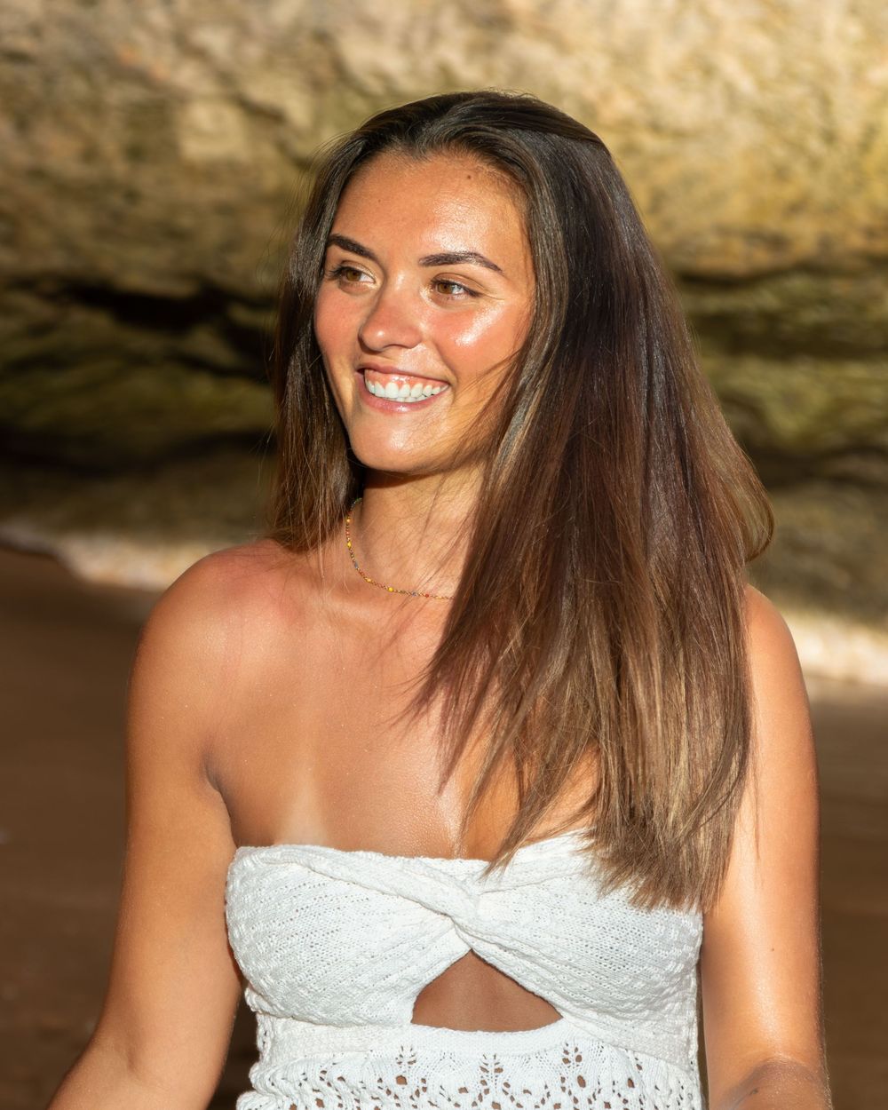 Smiling young woman with long brown hair wearing a white strapless top at a beach.