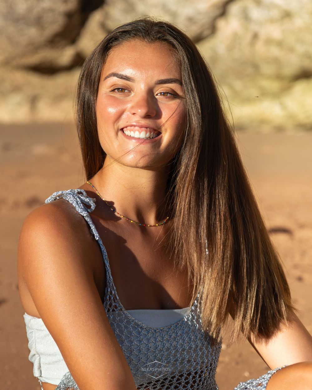 Young woman with long brown hair smiling outdoors at a rocky beach in a light blue crochet top.