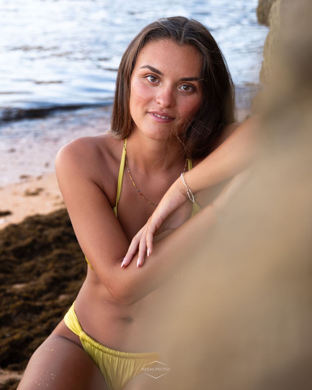 Young woman in a yellow bikini posing near rocky shoreline at the beach with calm water in background.