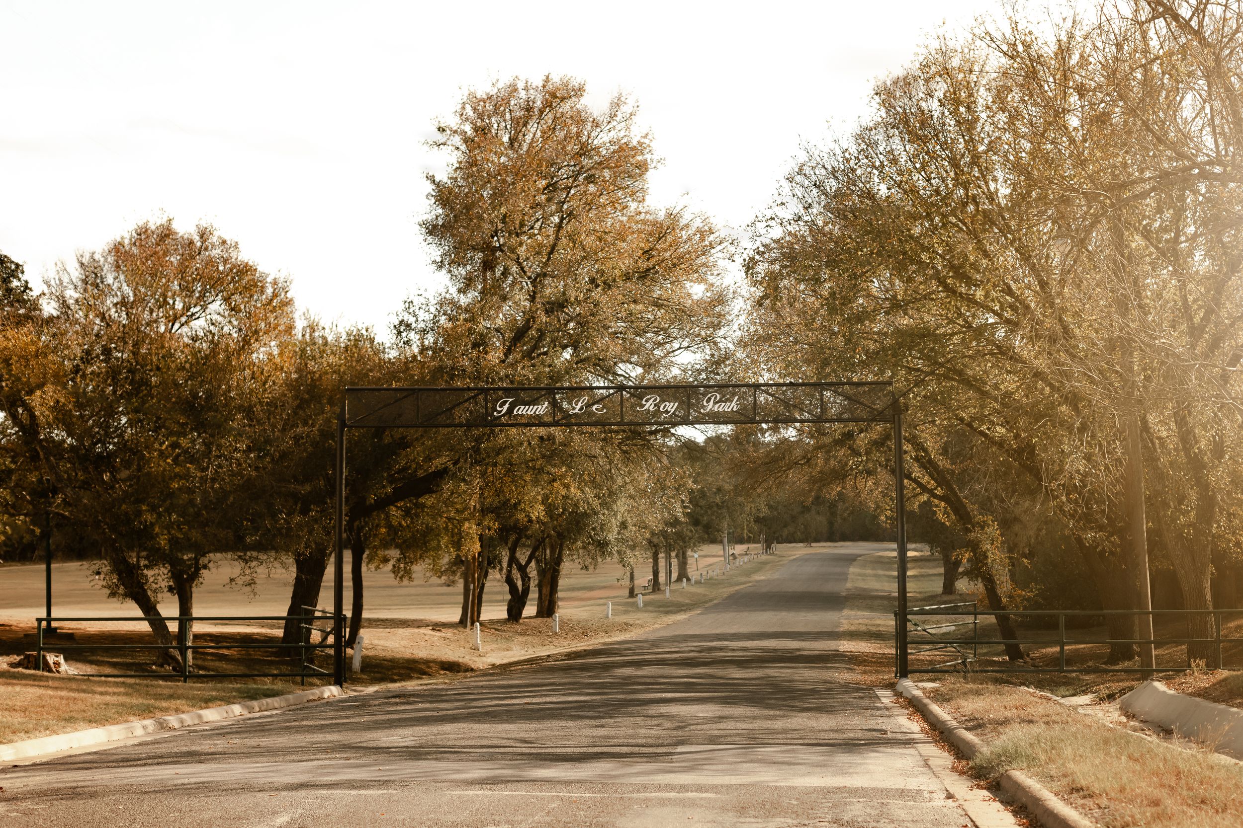 A sunlit road passes under a metal arch gate surrounded by autumn trees in a warm, golden landscape.