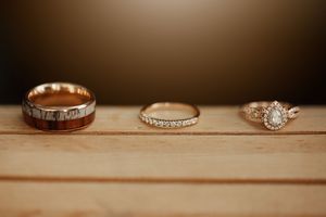 Three wedding rings on wood surface: rustic wood-inlay band, delicate gold band, and pear diamond halo engagement ring.
