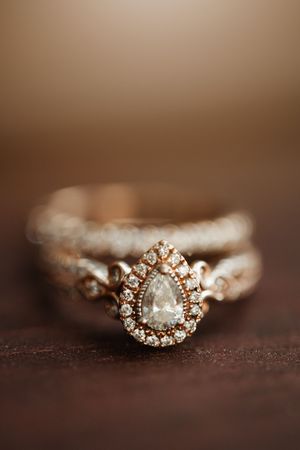 Close-up of a rose gold halo engagement ring with pear-shaped diamond and pavé band on dark surface.