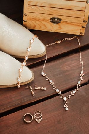 Bridal accessories flat lay with white cowboy boots, gold necklace, earrings, and rings on wood.