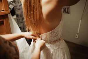 Bridesmaid fastening buttons on back of white lace wedding dress with gold belt detail.