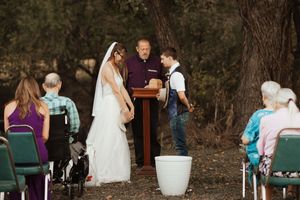 Outdoor wedding ceremony with bride in white gown and veil, young boy in suspenders, and officiant at rustic woodland altar.