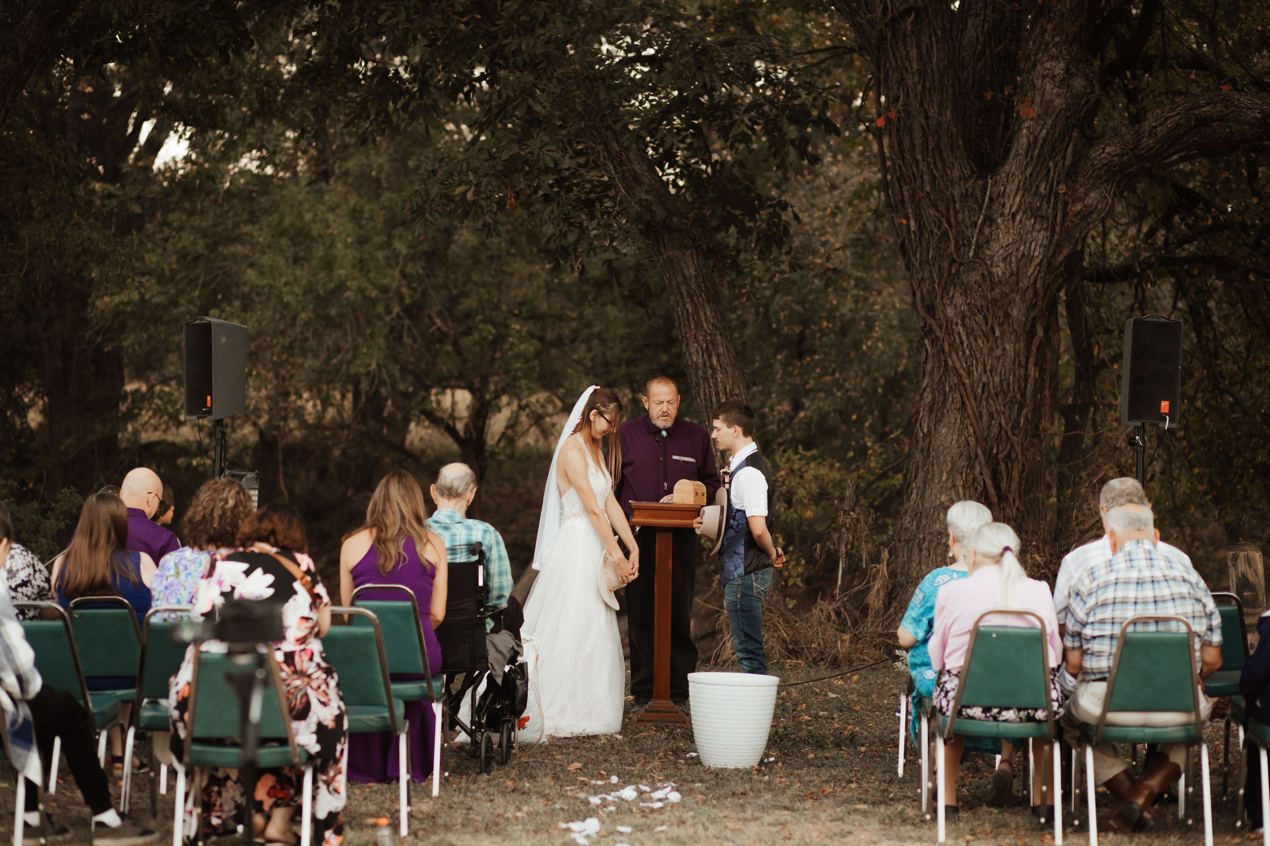 An outdoor wedding ceremony under large oak trees with guests seated in green chairs.