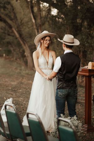 Bride in white lace gown and cowboy hat exchanges vows with partner outdoors in rustic woodland wedding ceremony.