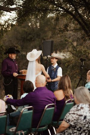 Outdoor country wedding ceremony with bride in white hat and groom in cowboy hat exchanging vows.