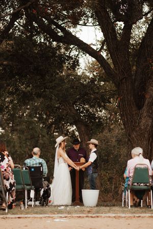 A rustic outdoor wedding ceremony with bride and groom in cowboy hats under a large oak tree.