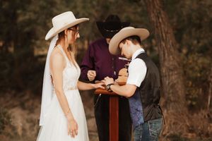 Bride and groom exchanging rings at rustic western wedding ceremony with officiant, both wearing cowboy hats outdoors.