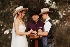 Western wedding ceremony with bride in white dress and groom in vest exchanging rings before officiant outdoors.