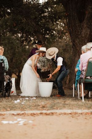 Bride and groom in cowboy hats performing a unity ceremony with a white bucket outdoors.