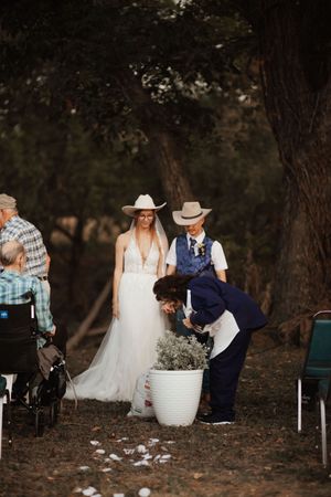 Bride and groom in cowboy hats stand outdoors at a rustic woodland wedding ceremony with guests seated nearby.