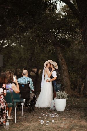 A bride and groom sharing their first kiss under oak trees at a rustic outdoor wedding ceremony.