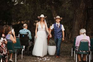 Bride and groom in cowboy hats walk down outdoor wedding aisle surrounded by seated guests among trees.