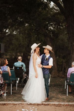 Bride and groom in cowboy hats share a romantic moment at an outdoor rustic wedding ceremony under large trees.