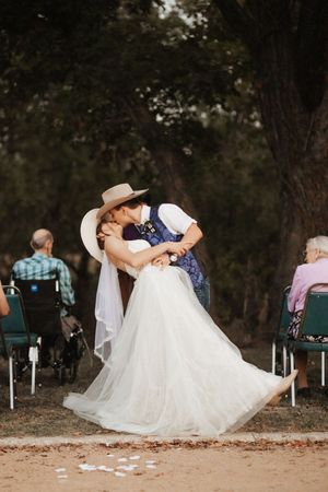 Bride and groom share a romantic dip kiss at outdoor wedding ceremony with guests seated among trees.