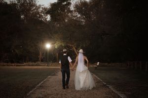 Bride and groom walking hand in hand down a tree-lined path at dusk in wedding attire.