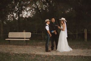 Outdoor rustic wedding ceremony with bride in white dress and groom in cowboy hat exchanging vows at dusk.