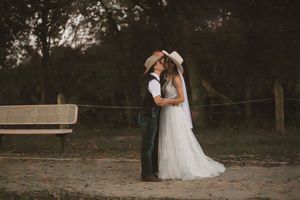 Bride and groom sharing a romantic kiss in cowboy hats, standing on a rustic dirt path surrounded by trees.
