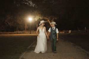 Bride in white gown and cowboy hat holds hands with groom in green suit and cowboy hat walking at night.