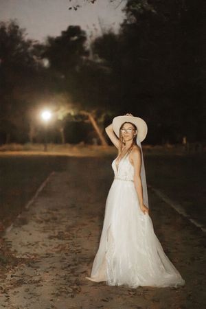 Woman in white tulle wedding gown and wide-brim hat poses on a path at night with a bright light glowing behind her.