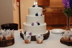 Three-tier white wedding cake adorned with purple and teal flowers on rustic wood slice display with floral arrangements.
