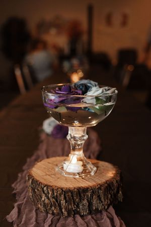 Crystal coupe glass with floating purple roses on rustic wood slice centerpiece at elegant event.
