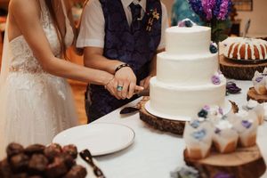 Bride and groom cutting a three-tier white wedding cake on a rustic wood stand at their reception.