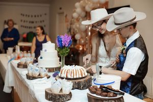 Two children in cowboy hats cutting cake at a rustic wedding dessert table with white tiered cake and bundt cake.