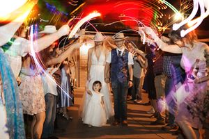 Bride and groom walk through sparkler tunnel with flower girl at outdoor wedding reception night celebration.