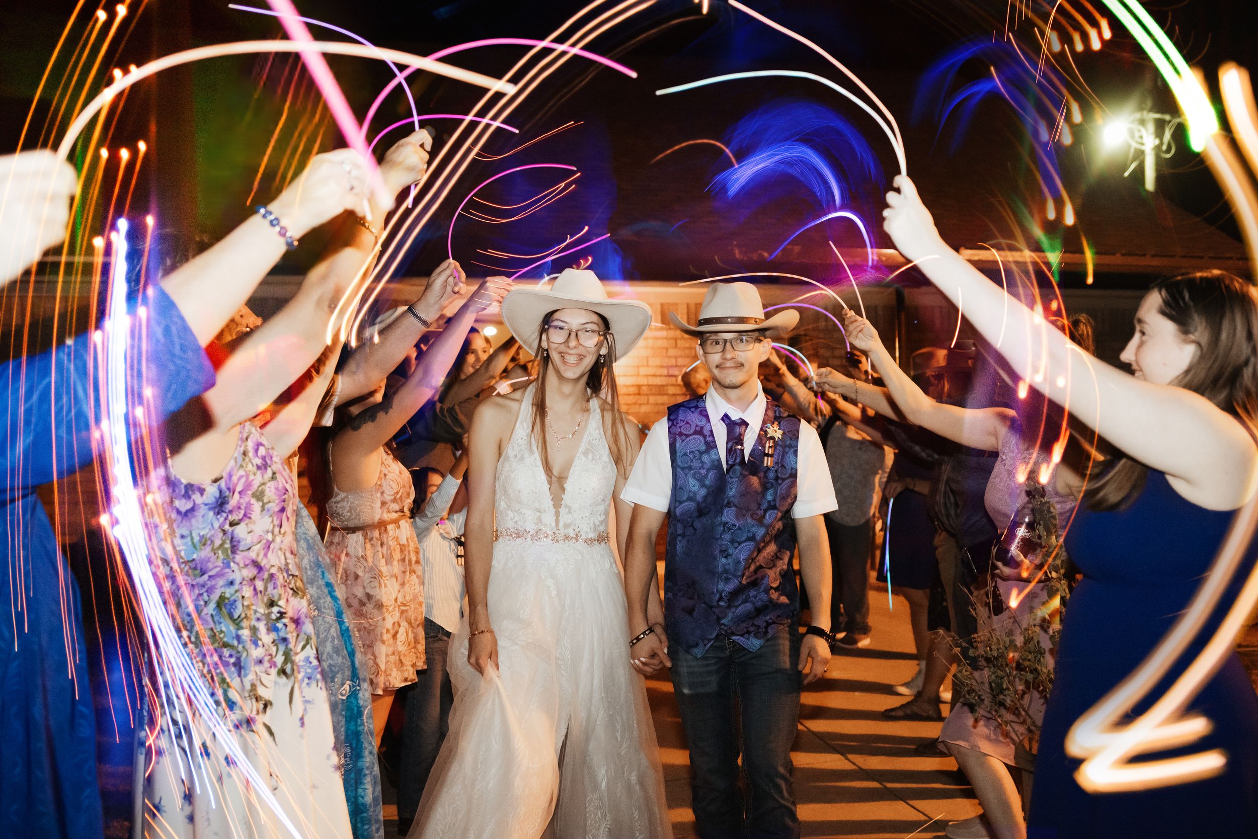 Bride and groom walk through sparkler tunnel surrounded by cheering guests at outdoor wedding reception at night.